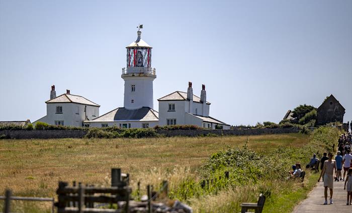 Caldey Island Lighthouse surrounded by open countryside on a sunny day, with visitors walking along the nearby path.
